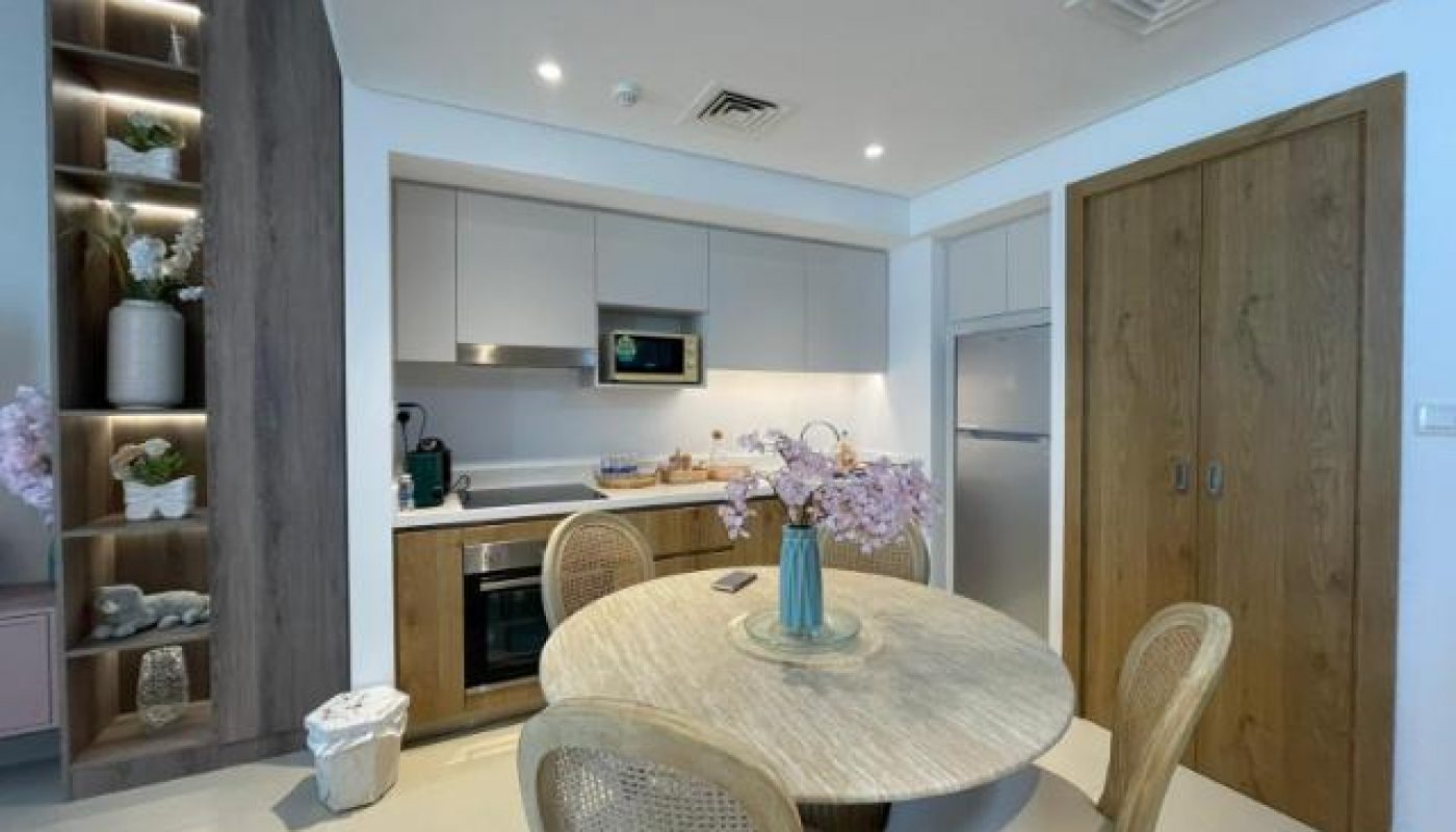 Modern kitchen and dining area with wooden accents, round table with four chairs, built-in shelves, and stainless steel appliances. Pink flowers in a vase decorate the table.