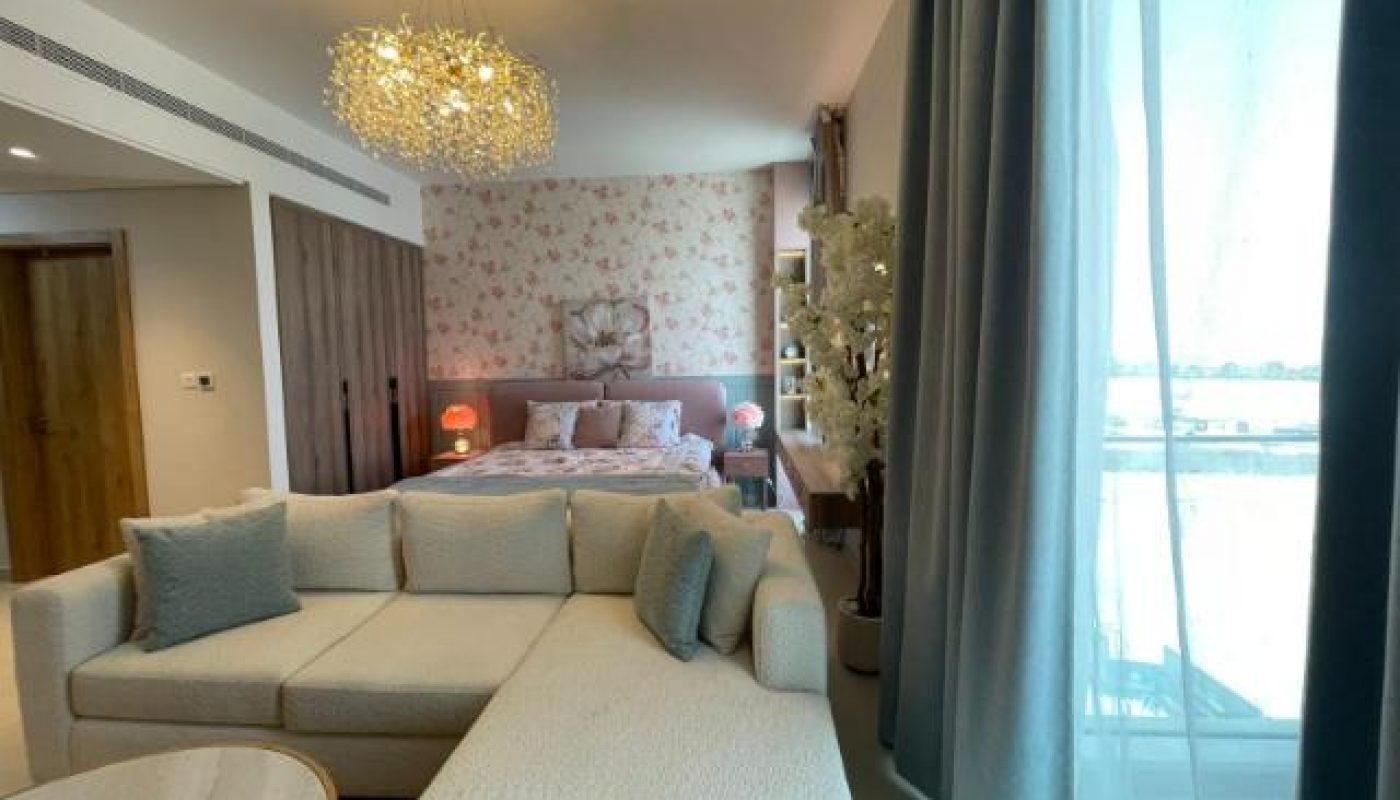 A bright, modern bedroom with a bed, floral wallpaper, a cream sofa, round table, decorative chandelier, and large window with sheer curtains.