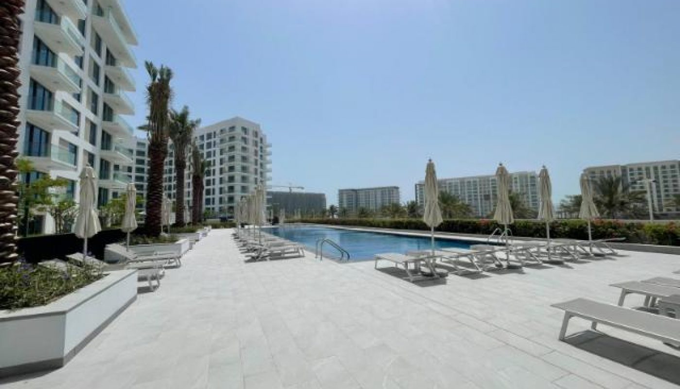 Outdoor swimming pool area with lounge chairs and umbrellas, surrounded by modern high-rise buildings and palm trees under a clear blue sky.