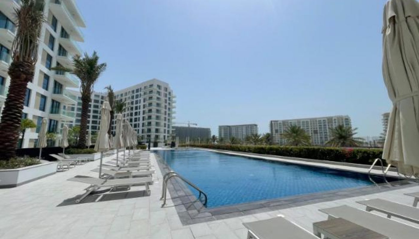Outdoor swimming pool surrounded by lounge chairs, umbrellas, palm trees, and modern apartment buildings under a clear blue sky.