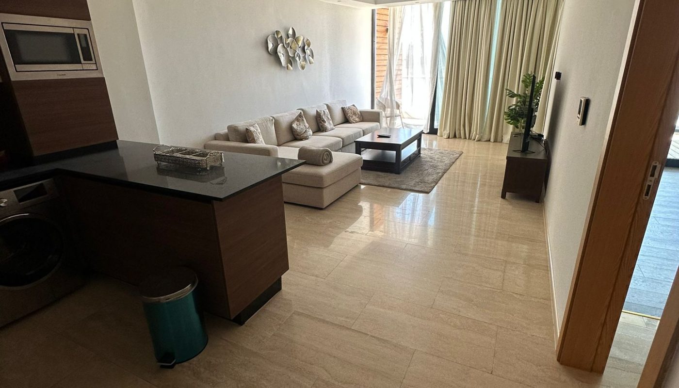 Modern living room with beige tiled floor, a light-colored sectional sofa, coffee table, wall art, and large windows with curtains; kitchen area visible on the left.