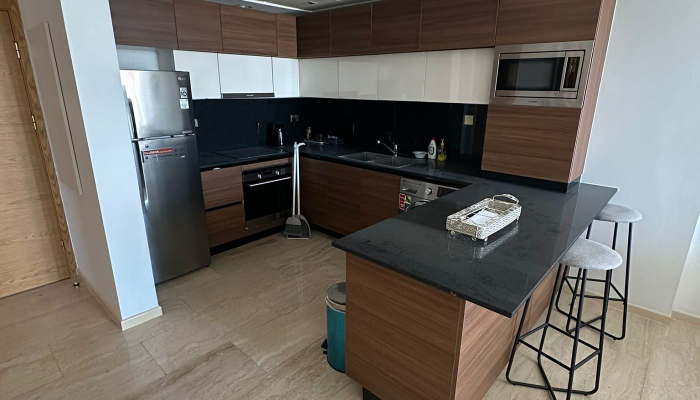 Modern kitchen with wood and black cabinets, stainless steel appliances, a black countertop island with two stools, and light beige tile floor. Cleaning supplies are visible on the floor.