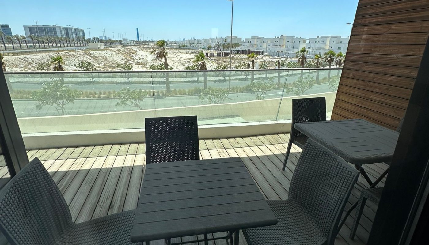 A balcony with two dark tables and several chairs overlooks a street and buildings under a clear blue sky.