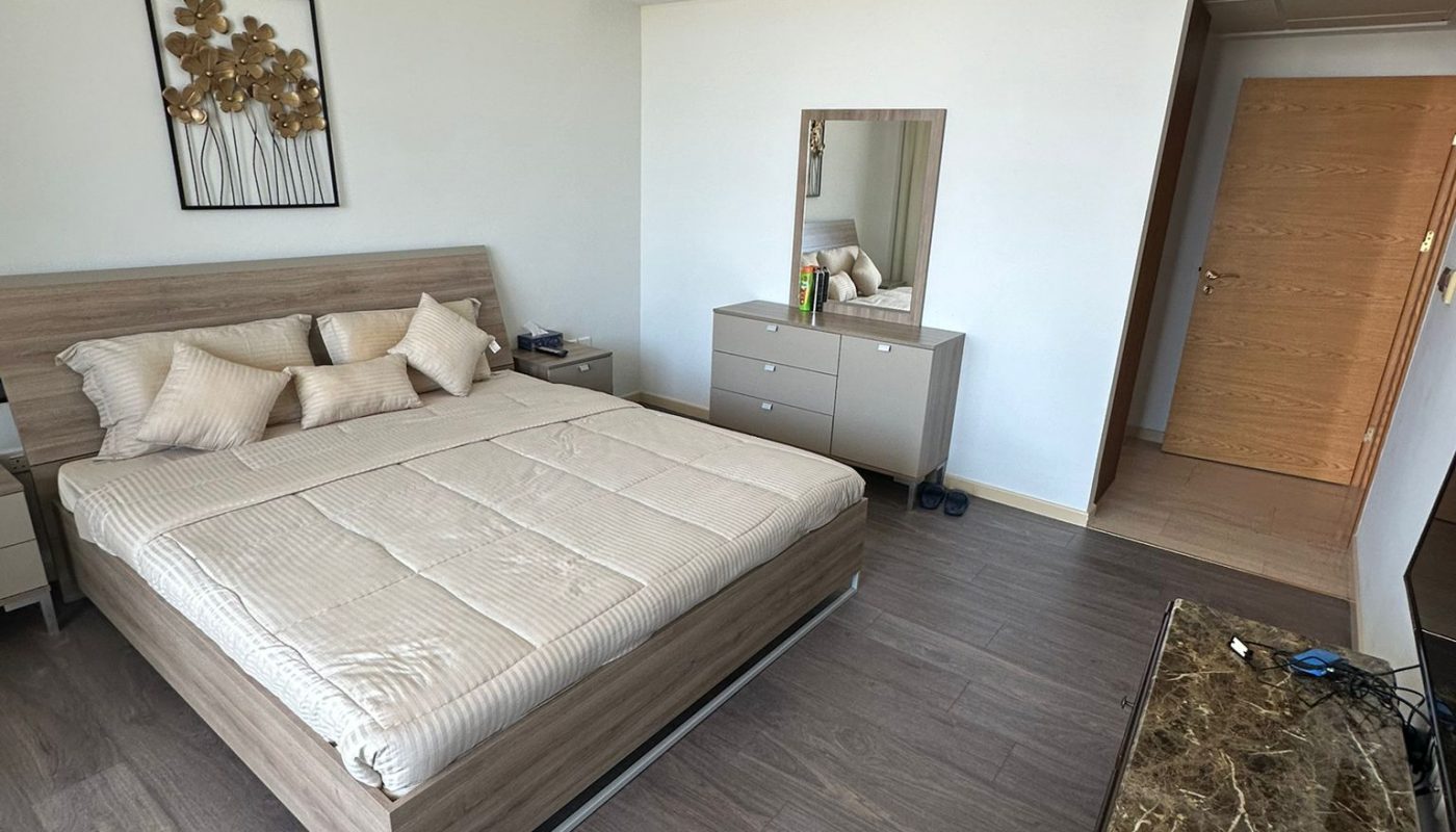 A modern bedroom with a made bed, light wood furniture, a marble-topped table, a dresser with a mirror, and a closed wooden door.