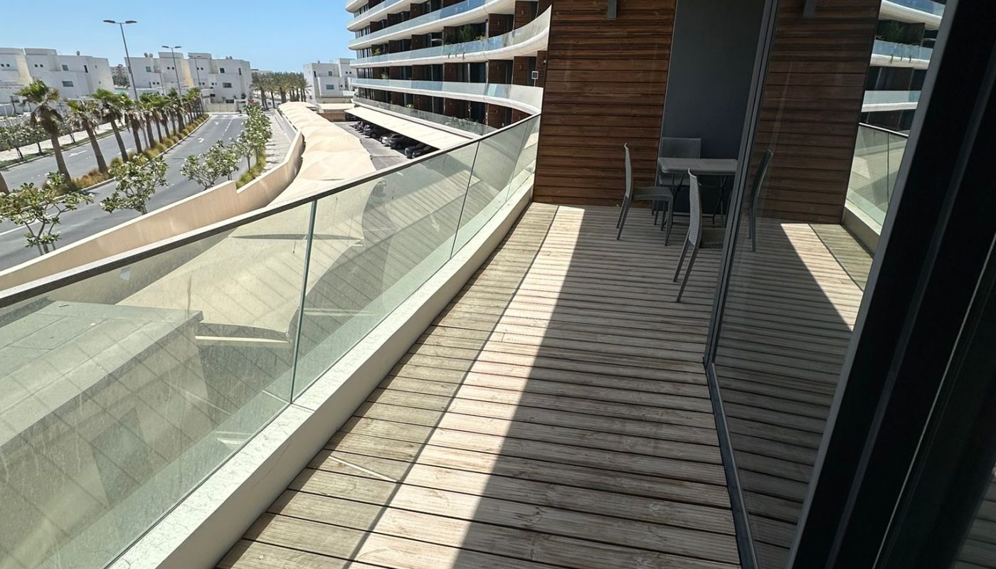 View of a modern apartment balcony with wooden decking, glass railing, and outdoor seating, overlooking a curved building and a street with trees under a clear blue sky.