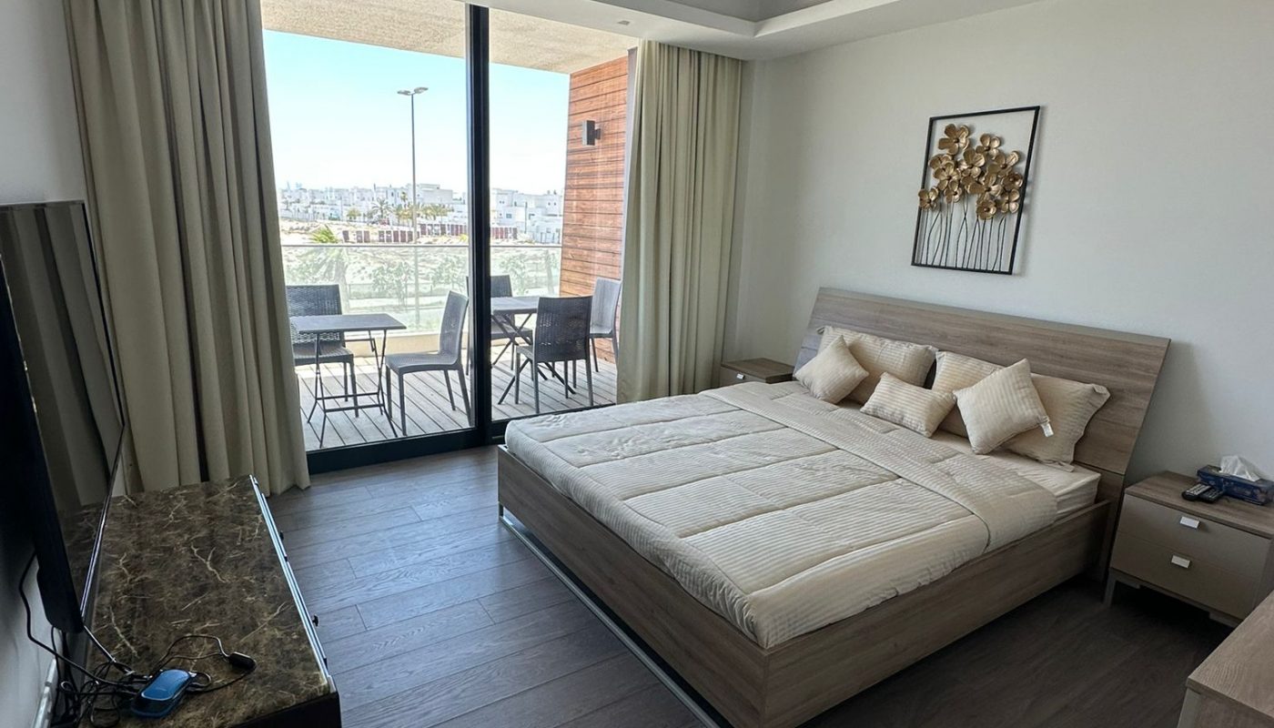 A modern bedroom with a double bed, TV, and nightstands, featuring a balcony with outdoor seating and a city view through large glass doors.