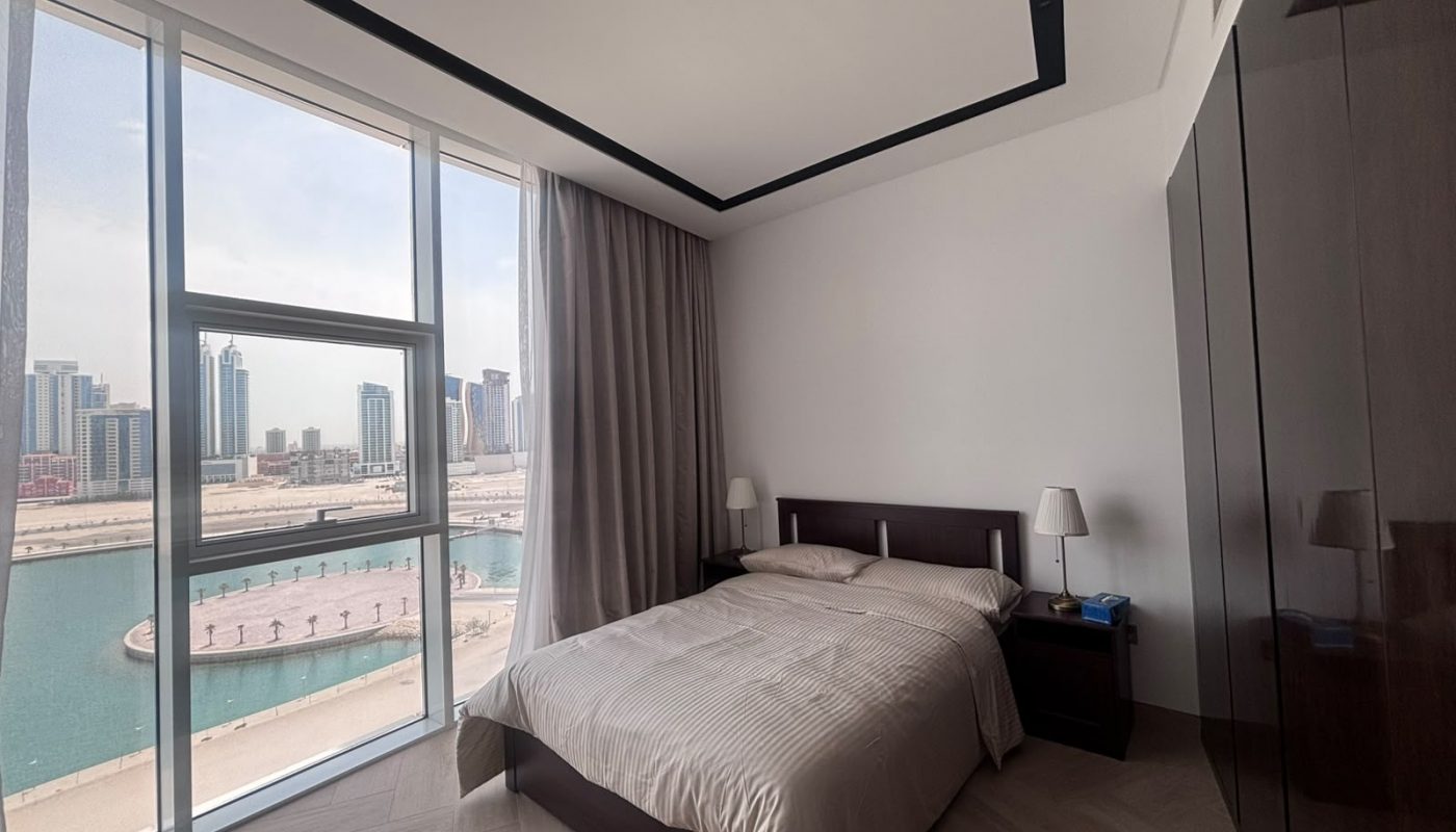 A modern bedroom with a double bed, two bedside tables, and large windows showing a city skyline and waterfront view.