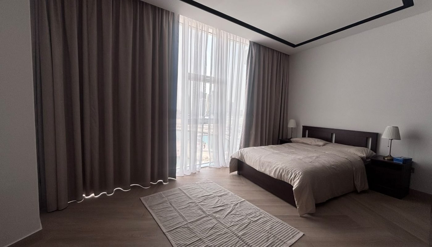 A minimal bedroom with a large window, beige curtains, a wooden bed with beige bedding, two nightstands with lamps, and a striped rug on a wooden floor.