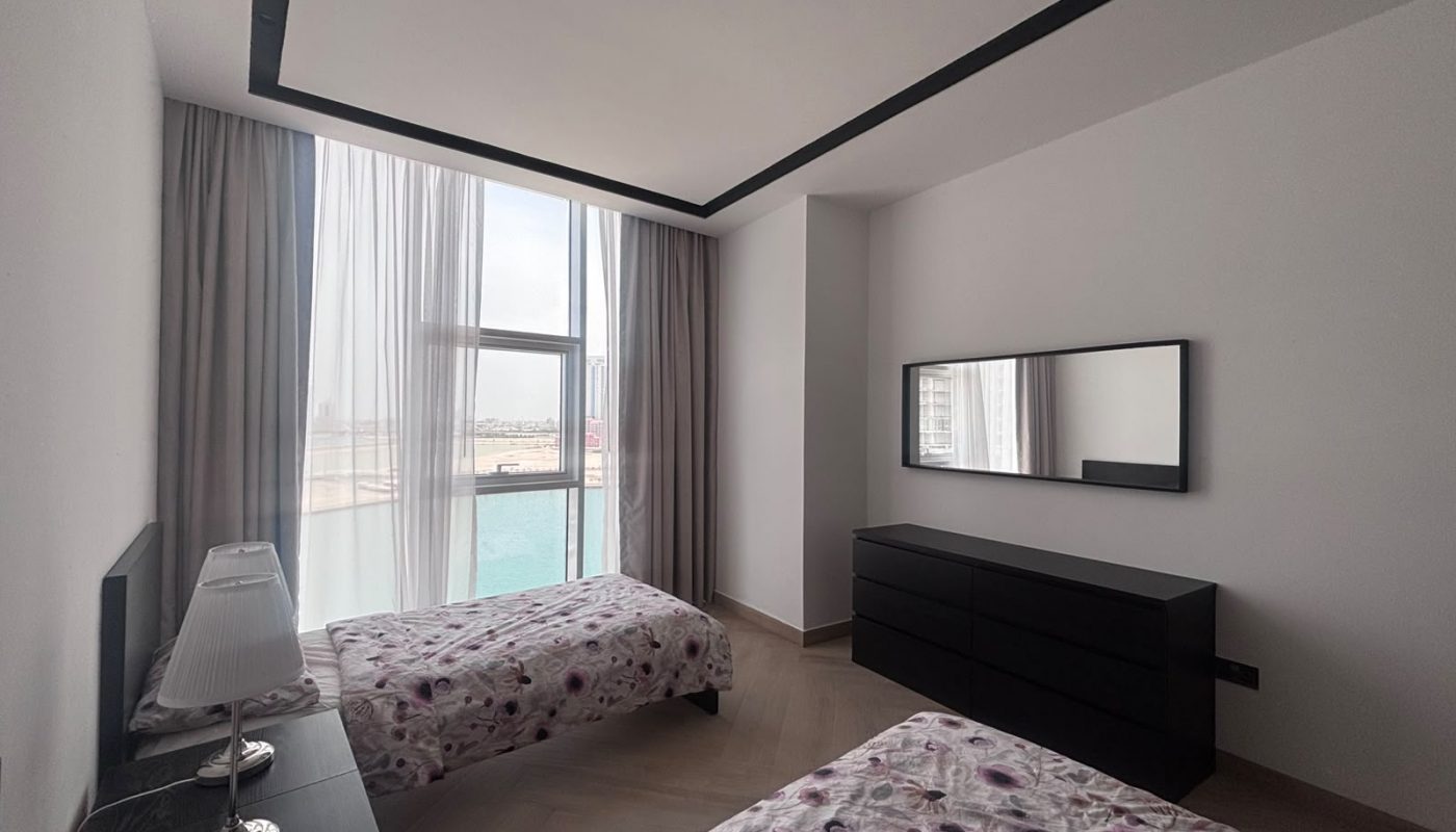 A modern bedroom with two single beds, floral bedding, a nightstand with a lamp, a dresser with a mirror, and large windows with sheer curtains overlooking a city and water.