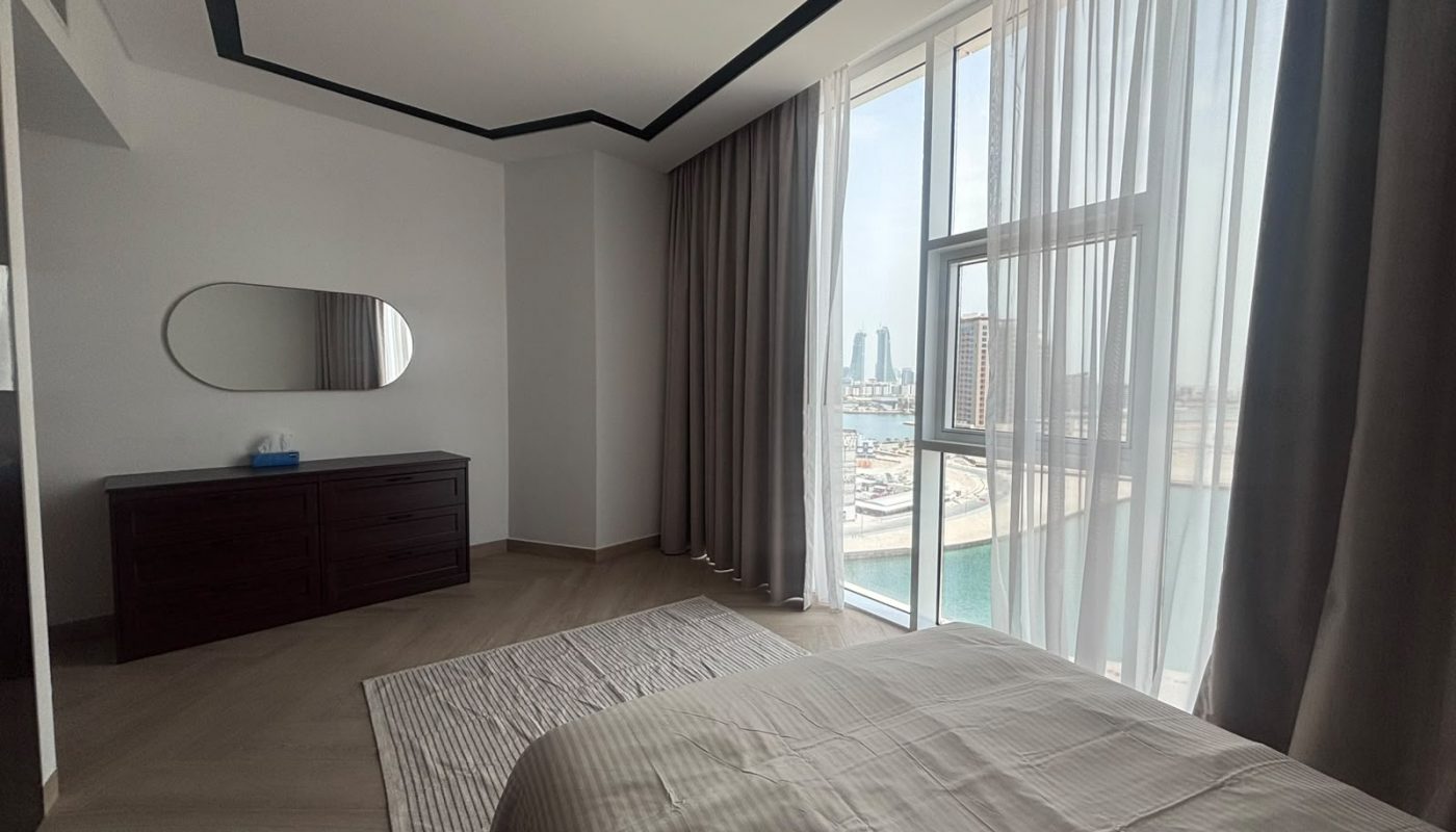 A modern bedroom with a bed, dresser, oval mirror, large windows with sheer curtains, and a view of city buildings and water outside.
