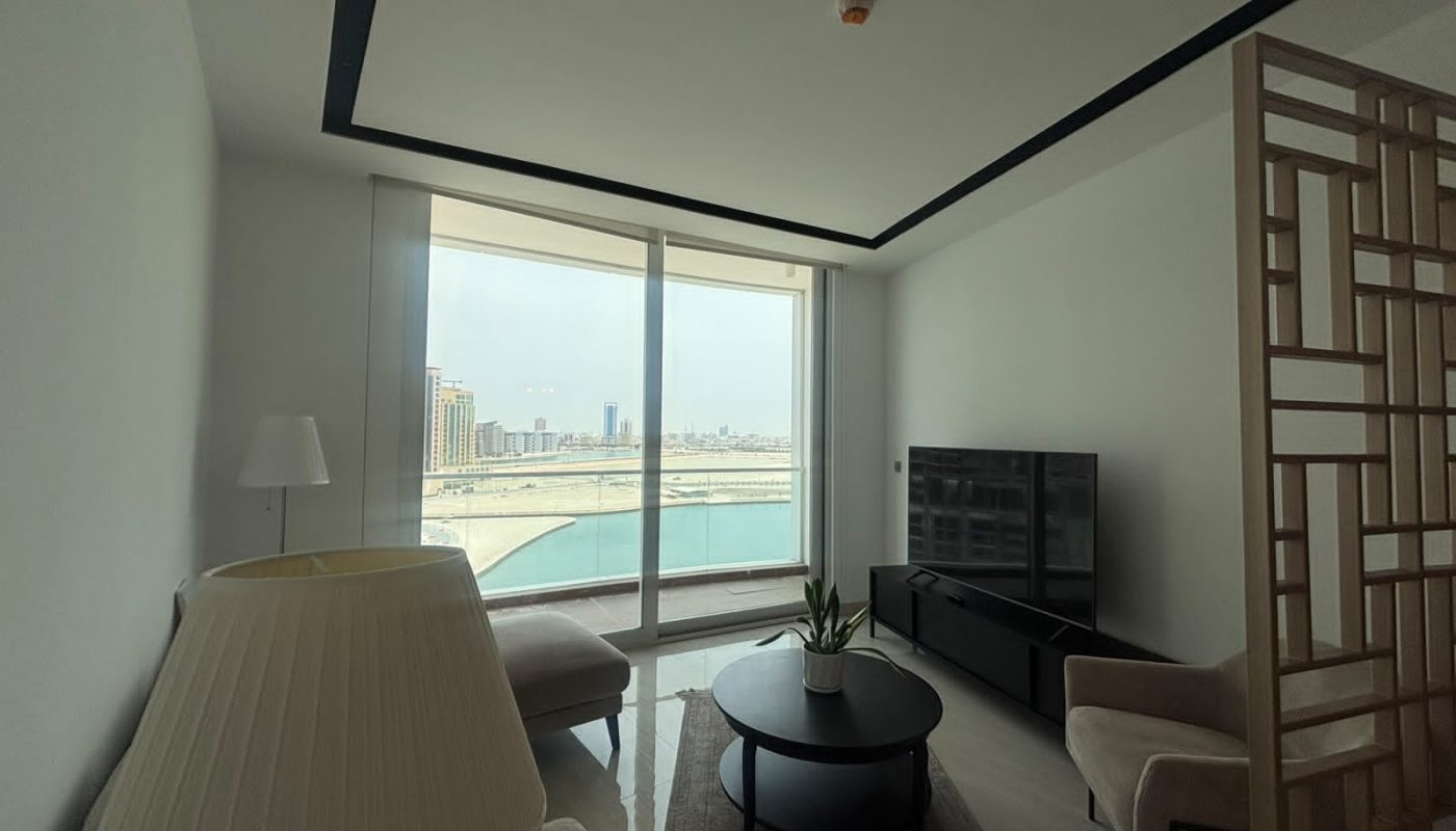 A modern living room with large windows overlooks a city skyline and water. The room has beige chairs, a round coffee table with a plant, and a TV on a black stand.