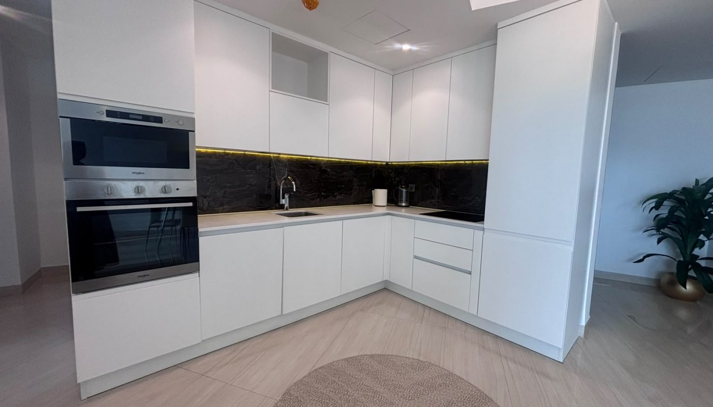 Modern white kitchen with built-in oven, stovetop, black backsplash, under-cabinet lighting, and a round rug on a light wood floor. A potted plant is visible at the right edge.