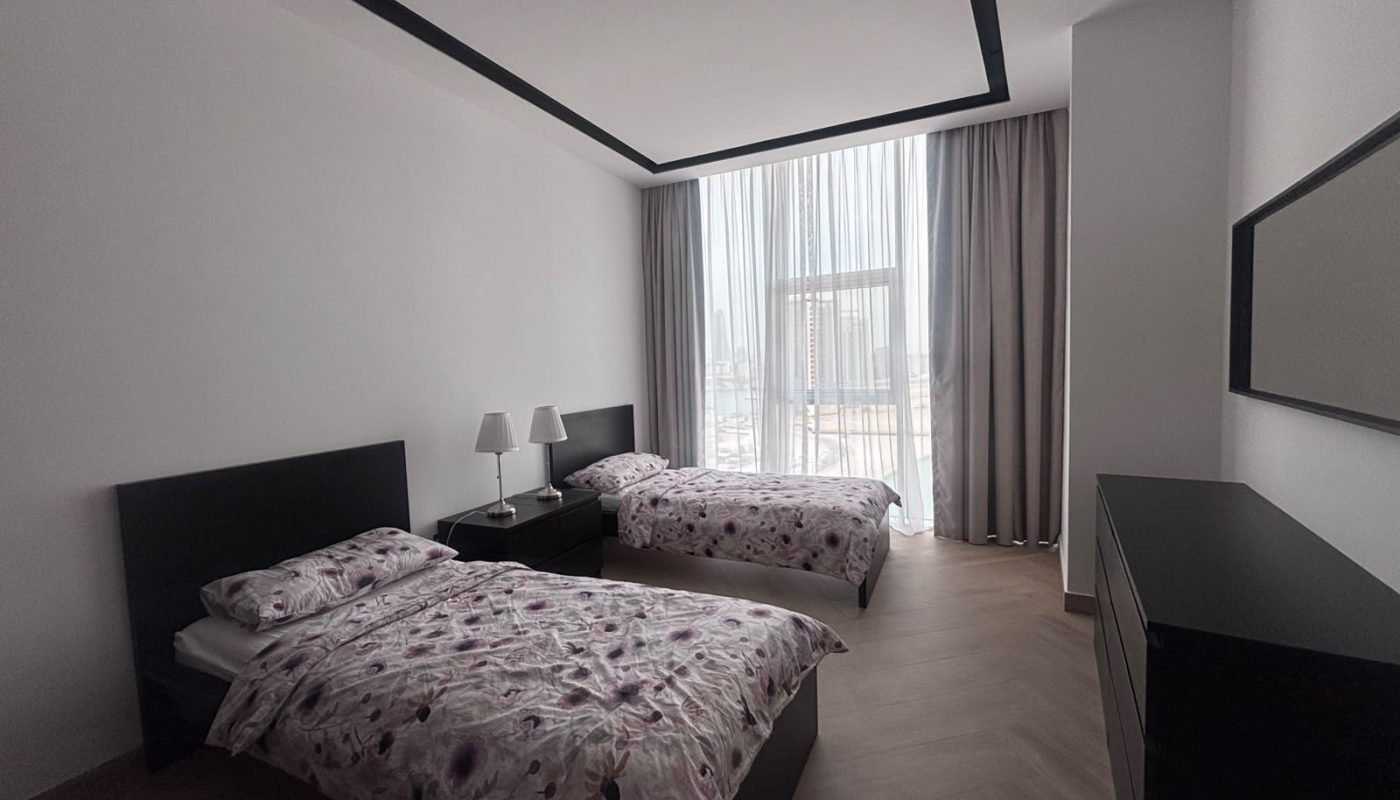 Minimalist bedroom with two single beds, floral bedding, a window with curtains, bedside table with two lamps, a dresser, and a wall mirror.