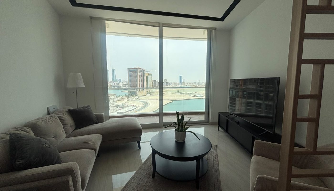 Modern living room with beige couches, a round coffee table, and a TV, featuring large windows overlooking a city skyline and waterfront.
