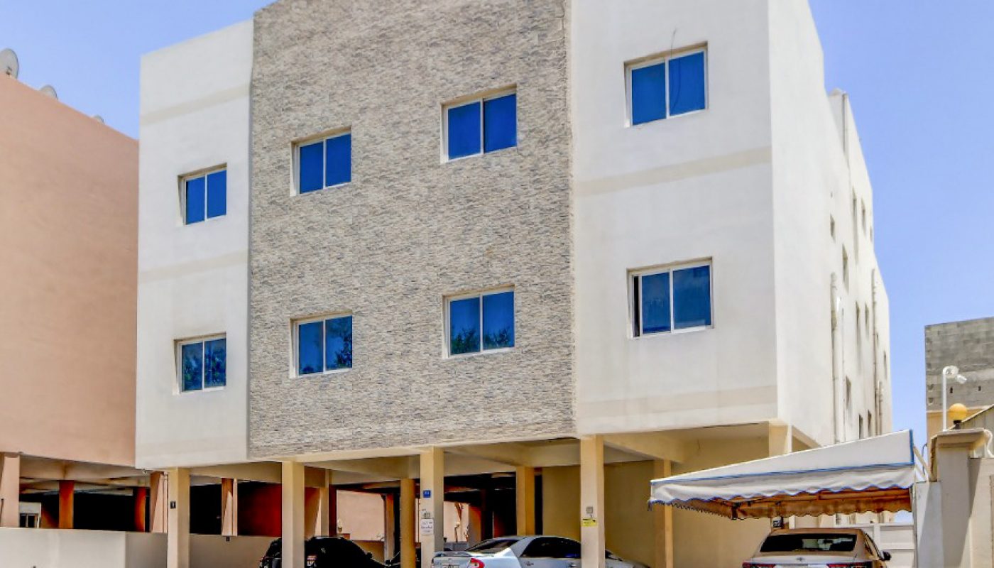 A three-story apartment building with a textured stone facade, several parked cars underneath, and a clear blue sky overhead.