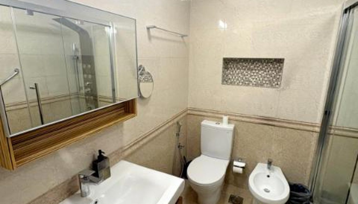 Modern bathroom with sink, mirror, toilet, bidet, shower enclosure, wall niche, and beige tiled walls and floor.