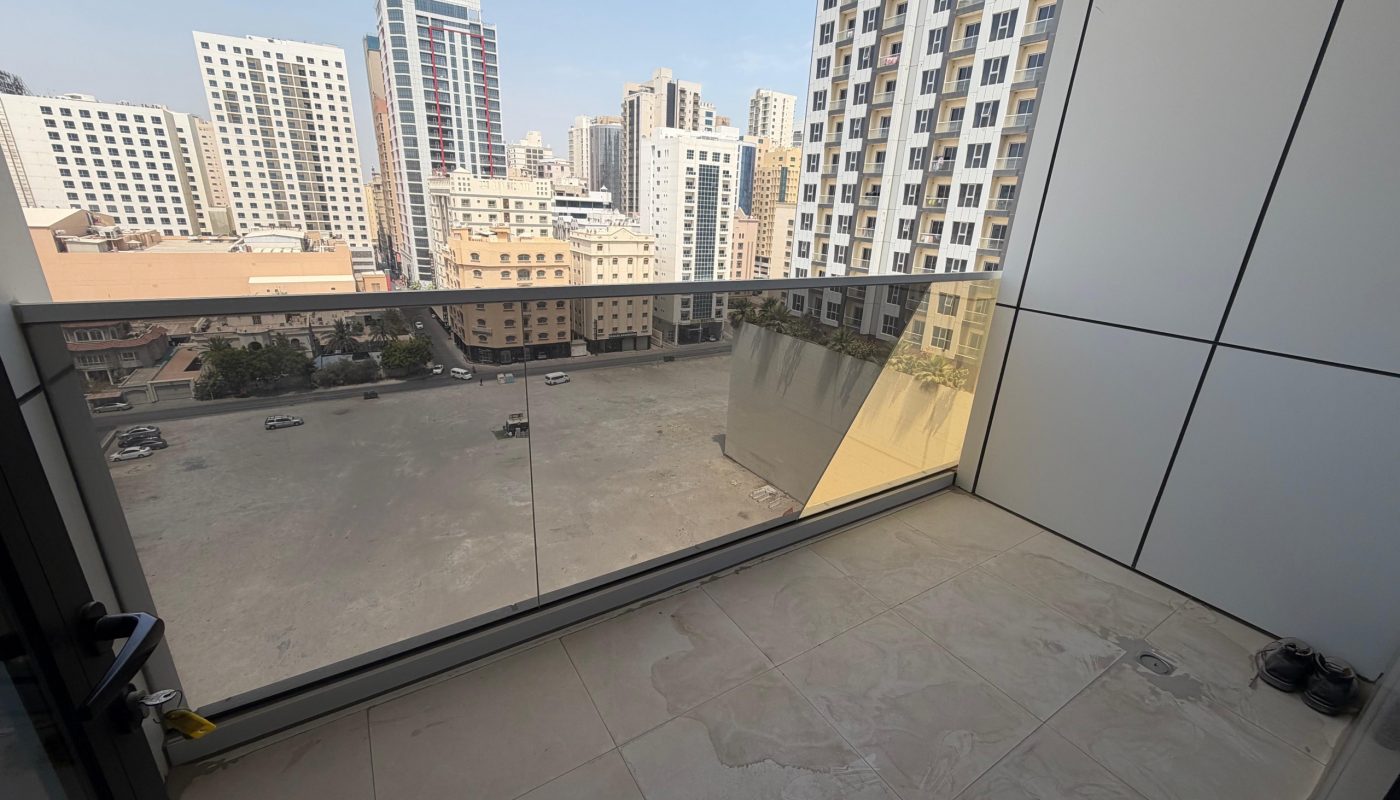View from a high-rise balcony with glass railing, overlooking a parking lot and multiple tall buildings on a clear day.