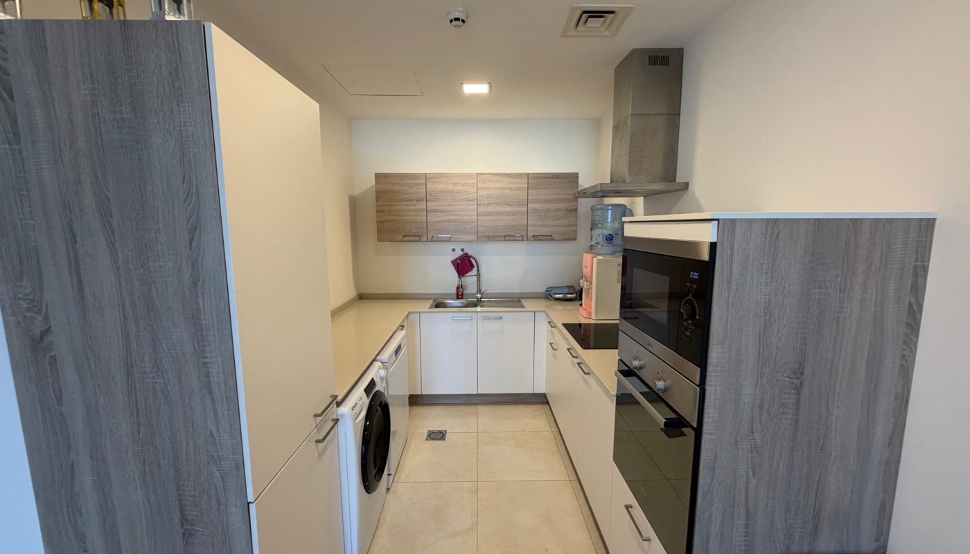 Modern galley kitchen with wood and white cabinets, built-in appliances, washing machine, and beige tiled floor. Countertop has a few small kitchen items and a water dispenser.