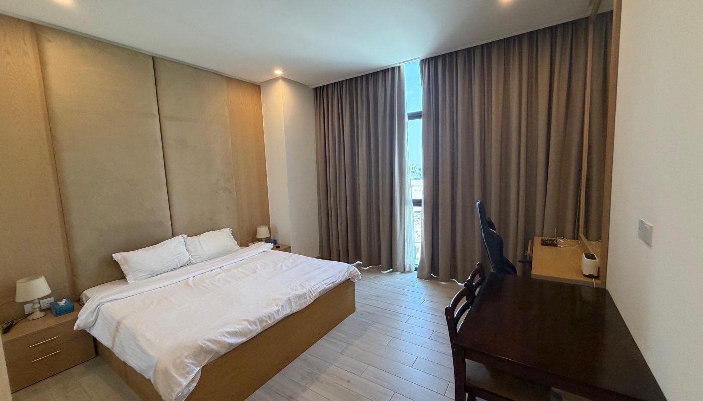 Modern bedroom with a large bed, side tables, a desk with a chair, and tall curtains covering a window.
