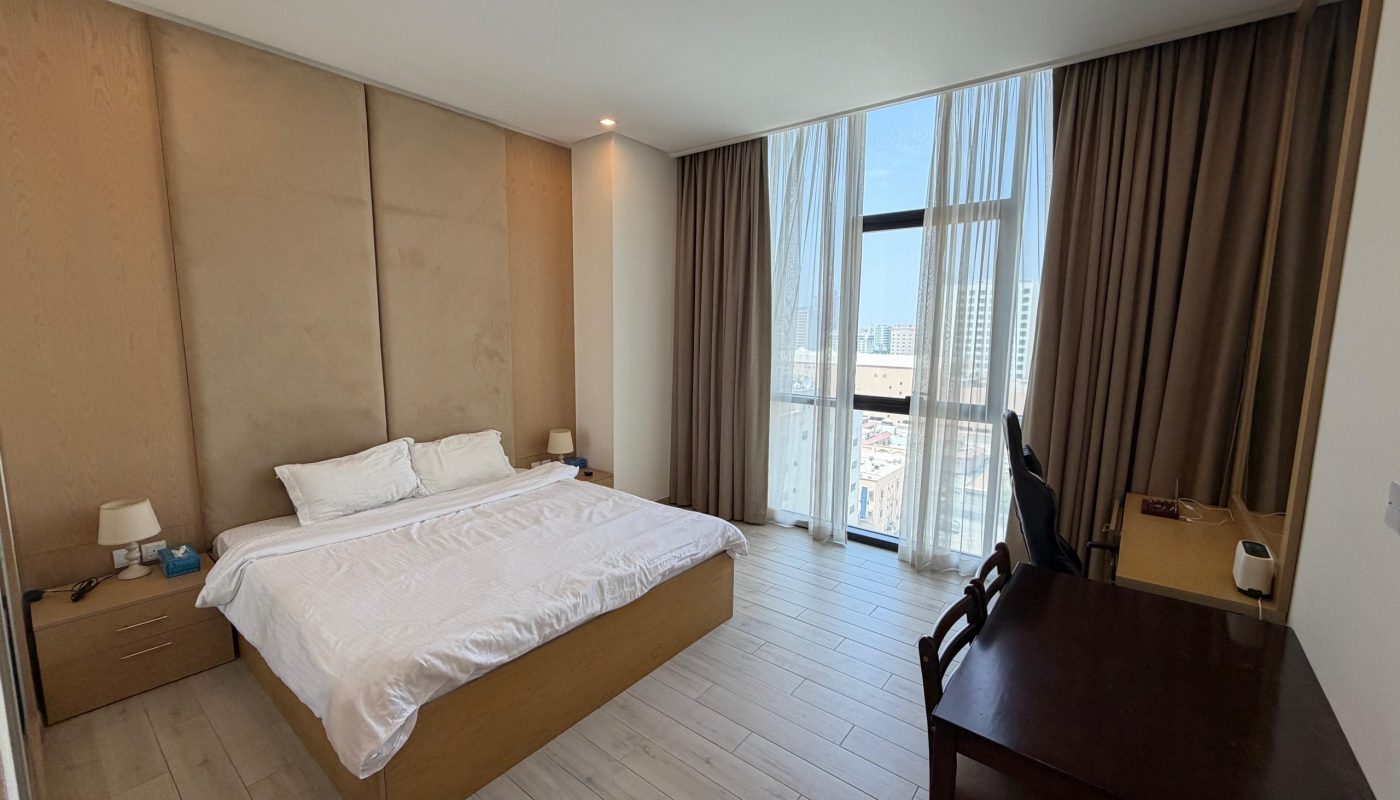 A modern bedroom with a double bed, two bedside tables, a desk with a chair, floor-to-ceiling window, brown curtains, and light wood flooring.