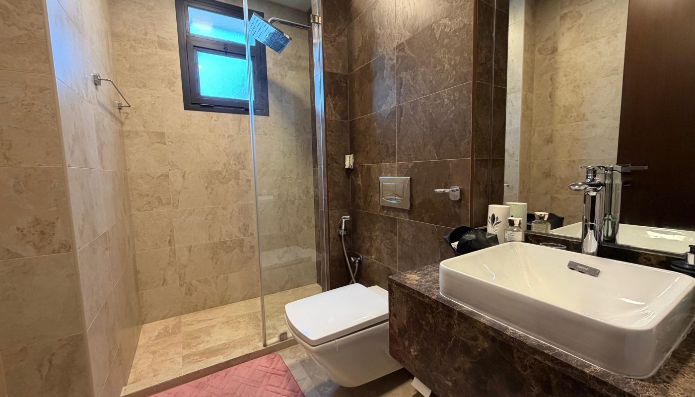 Modern bathroom with beige tiles, glass shower enclosure, wall-mounted toilet, white sink with chrome faucet, brown marble countertop, and a pink bath mat on the floor.