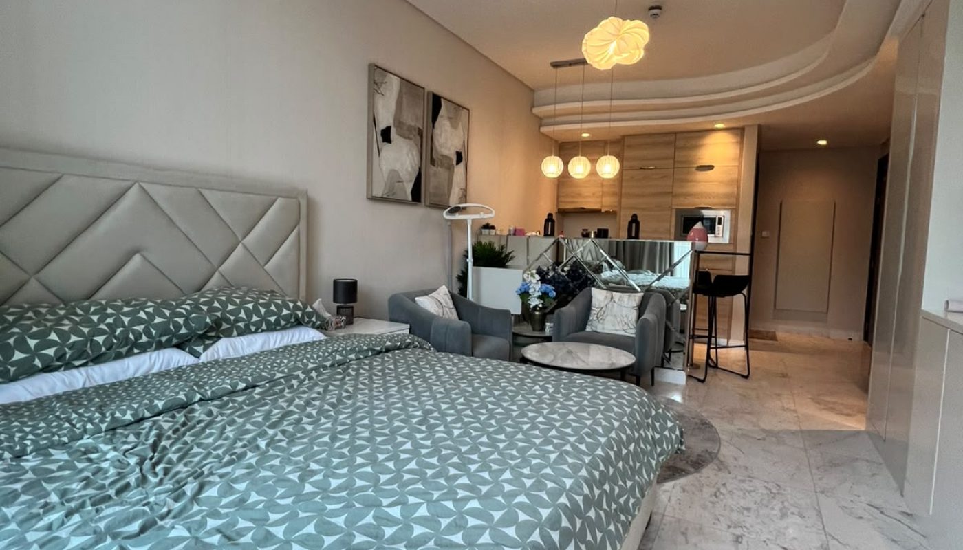 Modern studio apartment with a double bed, armchairs, small dining table, kitchenette, and marble flooring under soft lighting.
