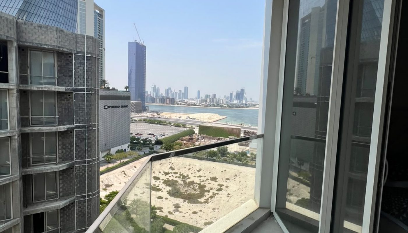 Glass balcony with a city skyline view, sandy lot below, tall buildings nearby, and clear blue sky overhead.