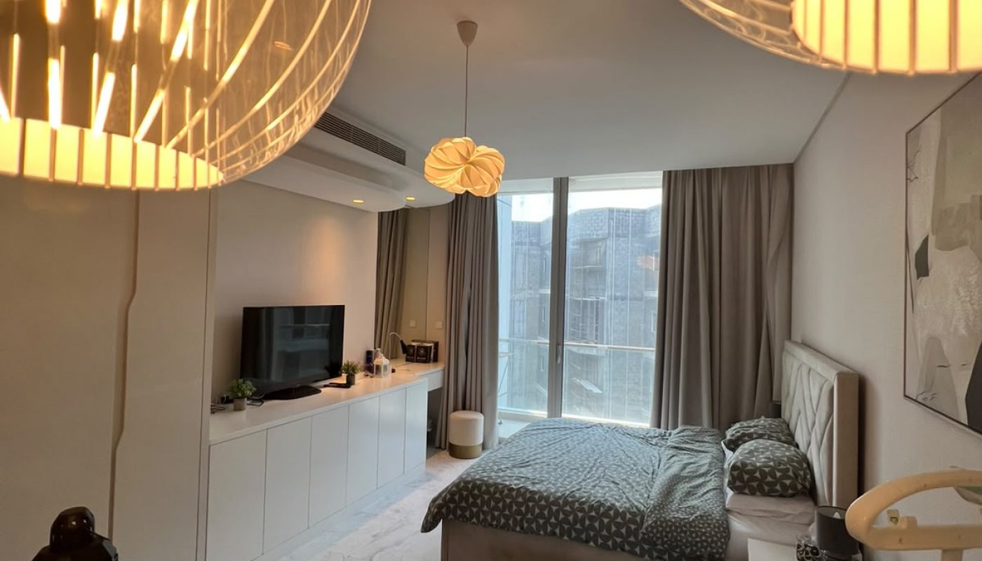 Modern studio apartment with a neatly made bed, gray sofa, TV, glass coffee table, large windows with curtains, and hanging pendant lights in the foreground.