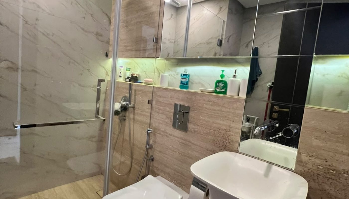 Modern bathroom with a glass-enclosed shower, wall-mounted toilet, pedestal sink, mirror cabinets, toiletries, and ambient green lighting above the mirror.