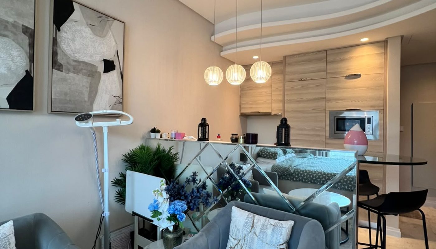Modern living room with two gray armchairs, abstract wall art, a mirrored console, kitchen area with wooden cabinets, and pendant lights. Blue flowers and decorative plants are visible.
