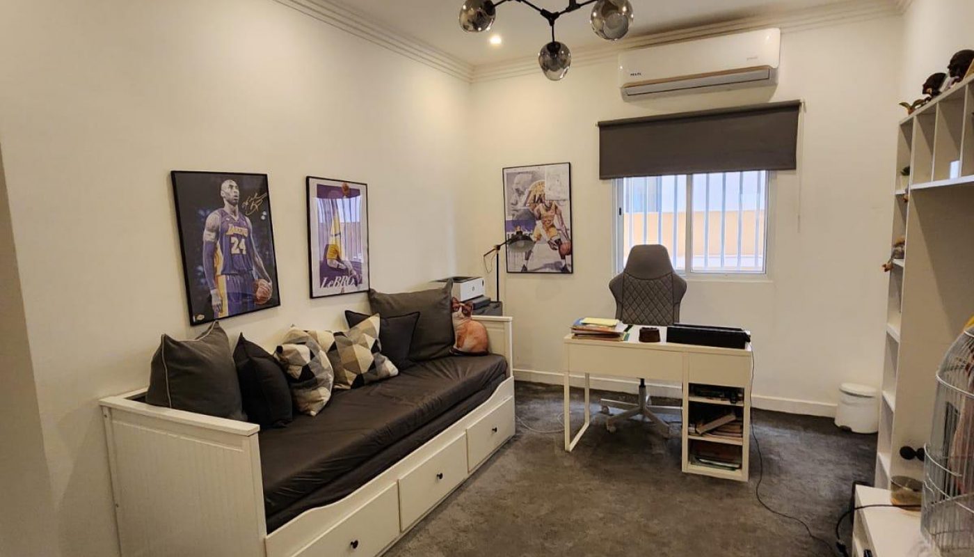 Home office with a desk, chair, framed sports posters, a sofa, shelves, air conditioner, and a window with blinds. Modern ceiling light fixture and gray carpet flooring.