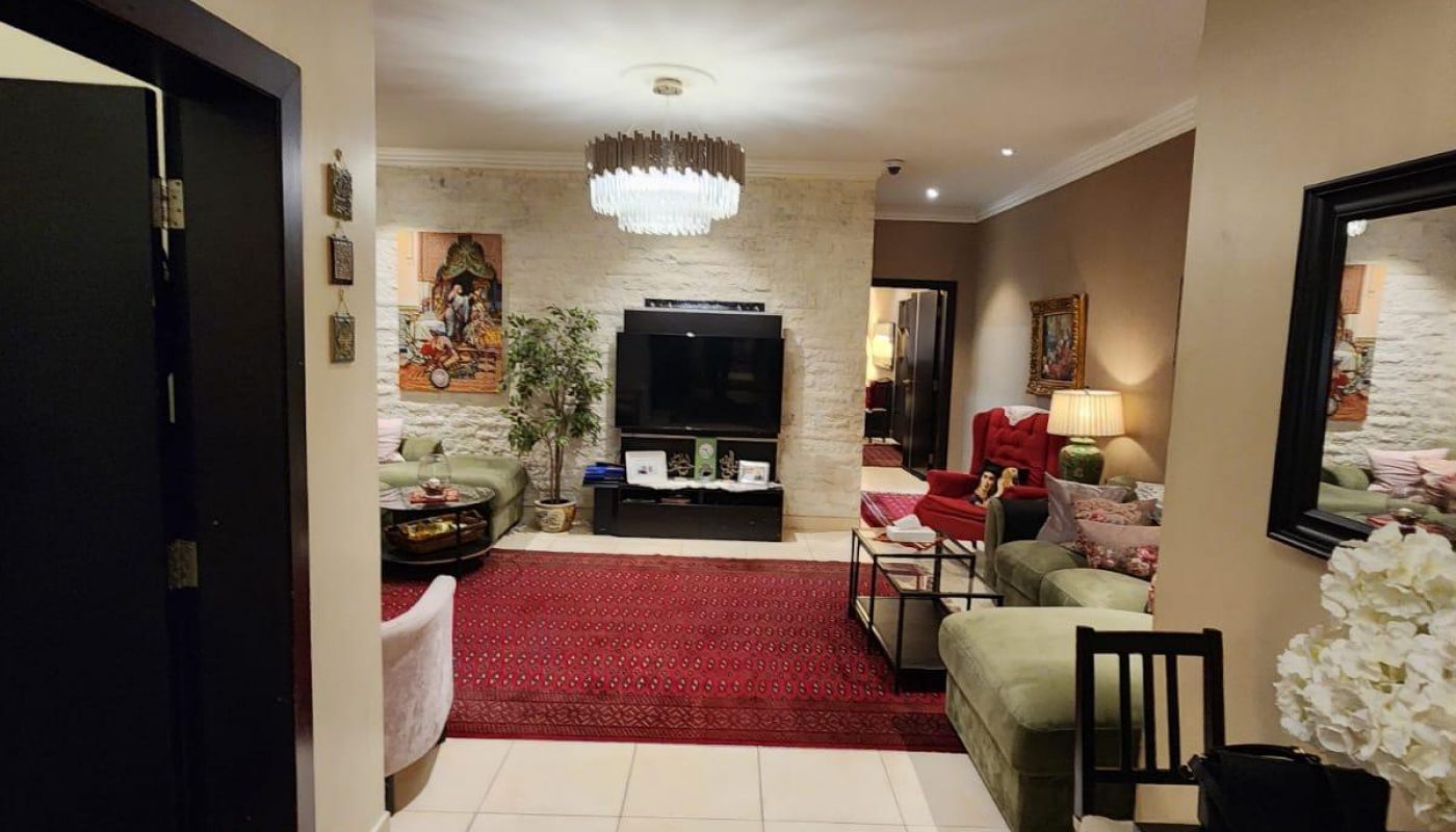View of a warmly lit living room with a TV, sofas, armchairs, rugs, wall art, and chandeliers, seen from a tiled entryway with a dark wooden console.