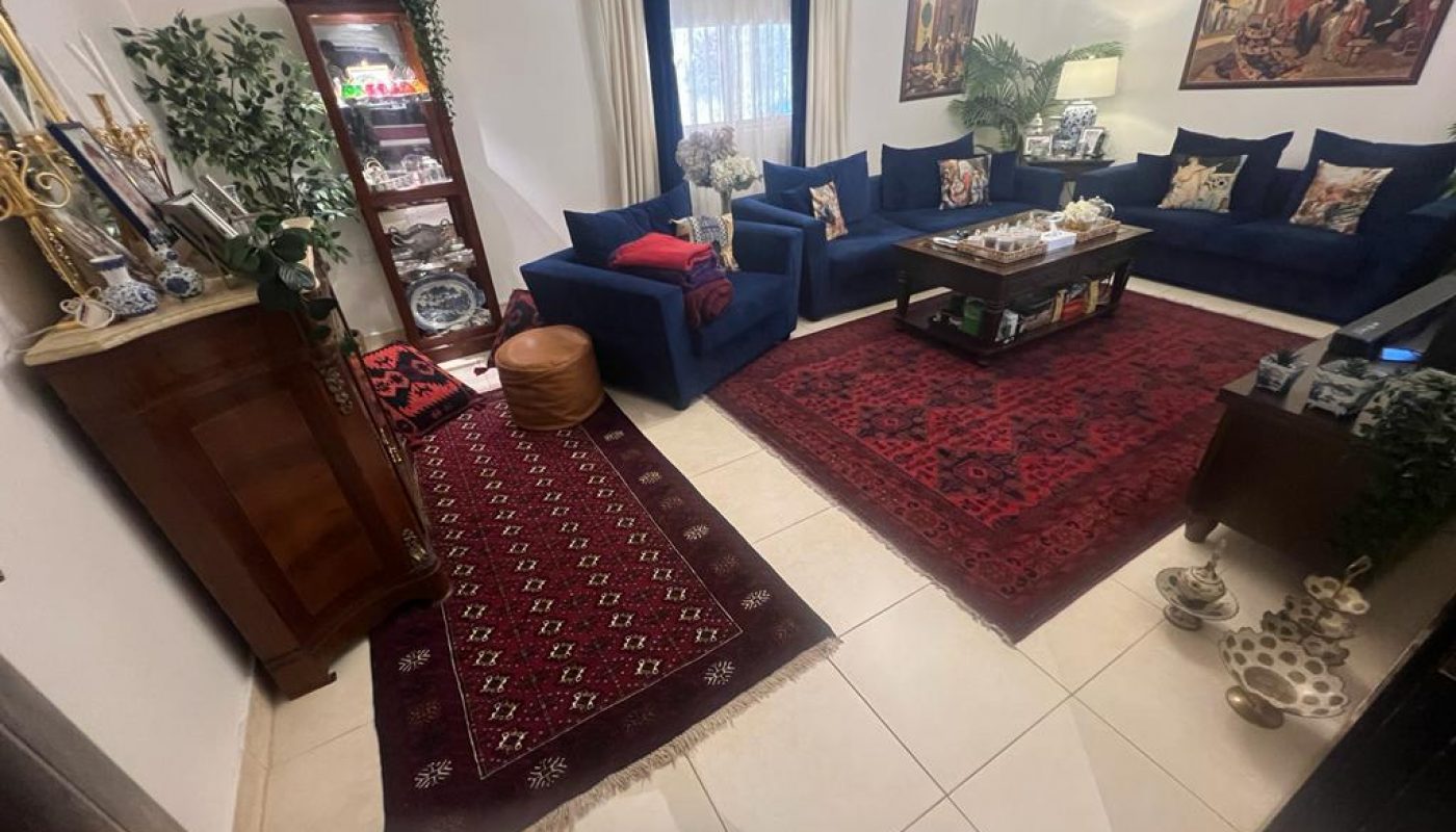 A living room with blue sofas, a red patterned rug, wooden cabinet, coffee table, houseplants, and framed art on white walls. A window with white curtains lets in natural light.
