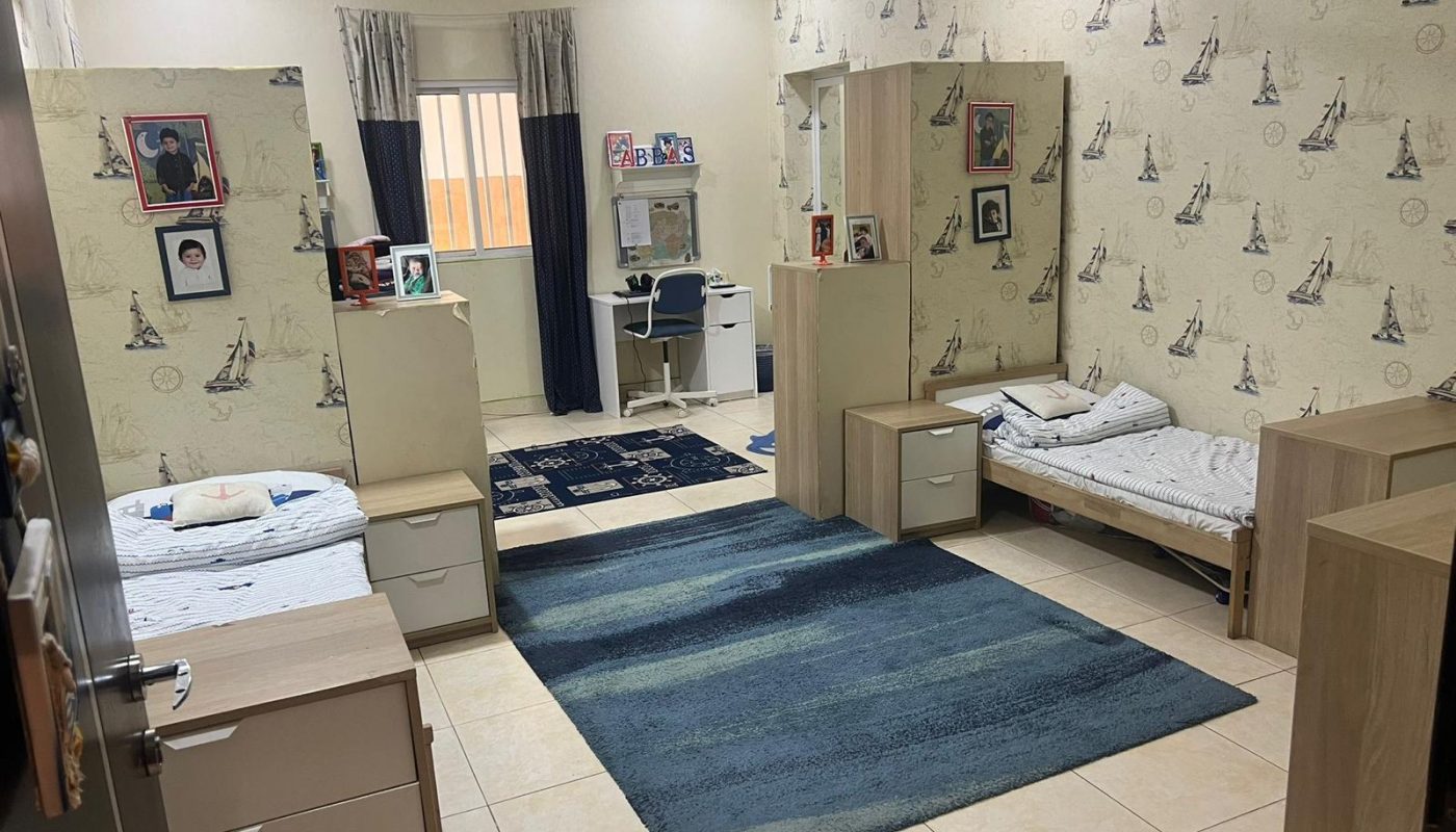 A tidy bedroom with two single beds, desks, bookshelves, and Eiffel Tower wallpaper. A window and a study area with a computer on a white desk are at the back.
