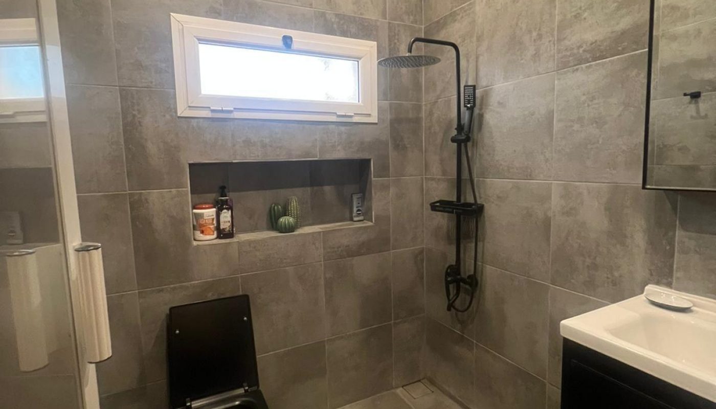 Modern bathroom with gray tiles, black toilet, wall-mounted shower, sink with mirror, small window, and built-in shelf holding toiletries and a small plant.