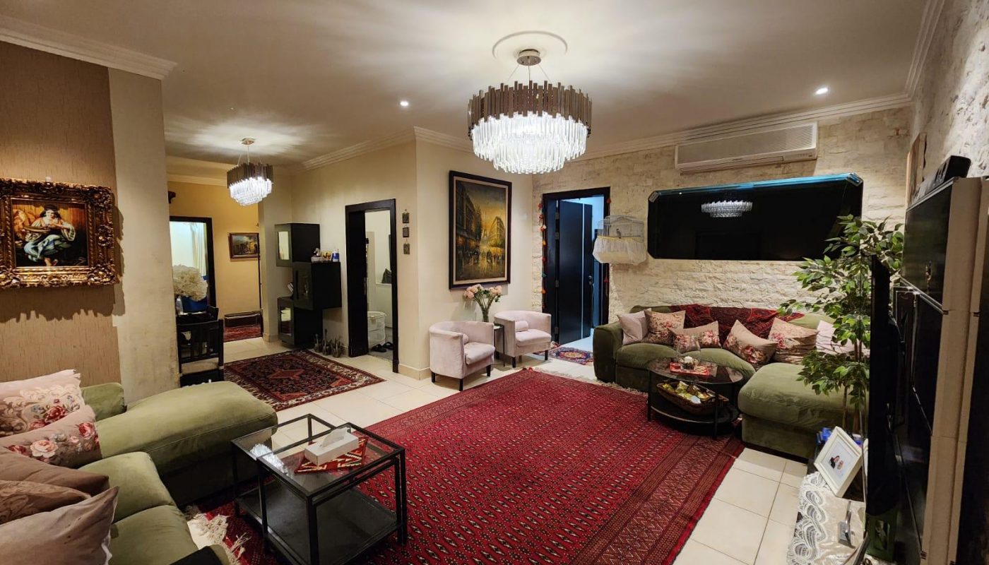Well-lit living room with green and pink sofas, glass coffee table, red patterned rug, chandeliers, and wall art; open door to balcony, and modern decor throughout.