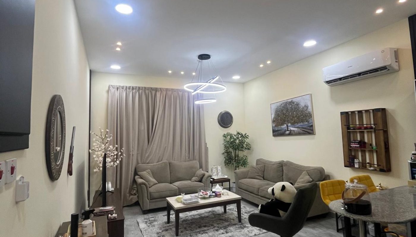 A modern living room with beige sofas, a geometric ceiling light, wall art, a dining table, and a split AC unit. The floor is tiled, and the room has decorative plants and various furniture.