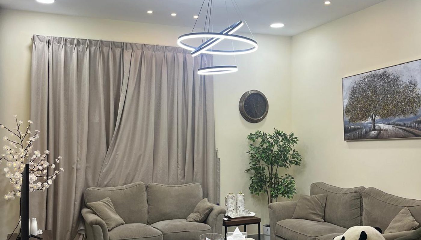 Modern living room with beige sofas, a coffee table, a green swivel chair with a panda plush, floor plant, wall art, and circular ceiling light. Neutral tones and minimalist decor.