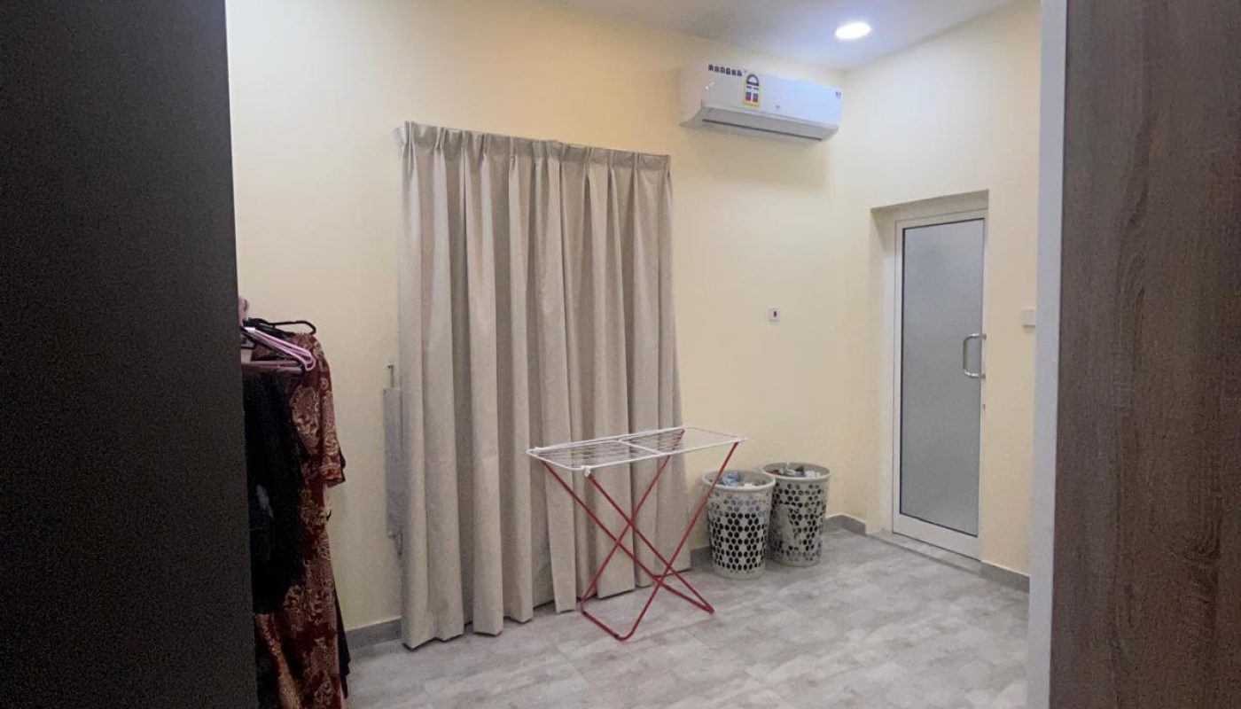 A simple room with gray tile floor, beige walls, closed curtains, a red drying rack, two patterned ottomans, a wall-mounted air conditioner, and a wardrobe.