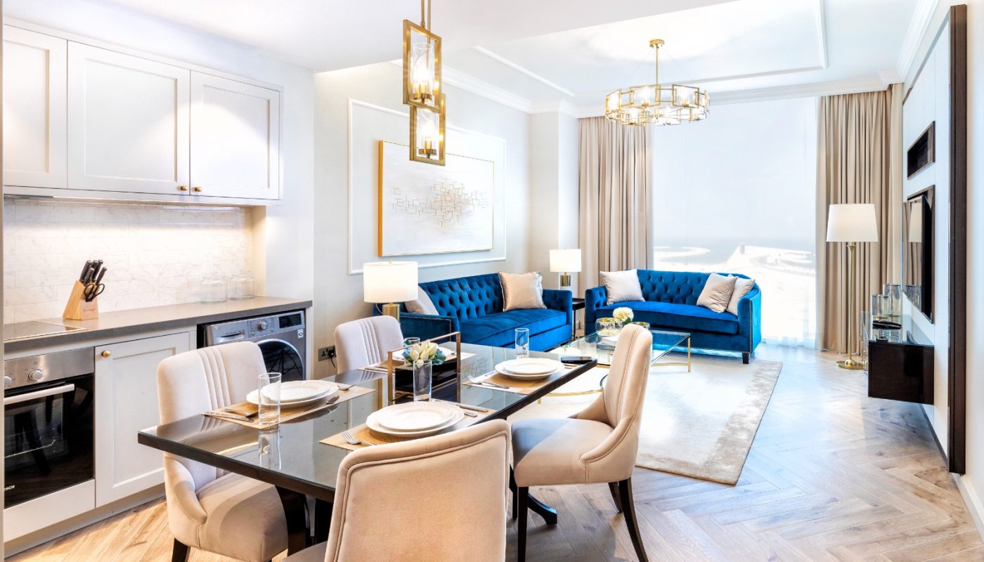 Modern open-plan living area with kitchen, dining table set for four, blue sofa, coffee table, and large windows with beige curtains, illuminated by gold pendant lights.