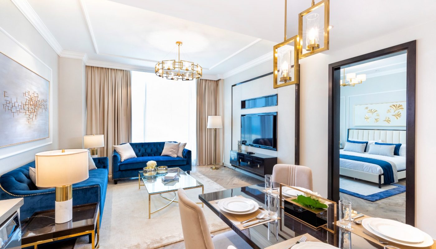 Bright modern living and dining area with blue sofas, a glass dining table set for four, and a visible bedroom in the background.
