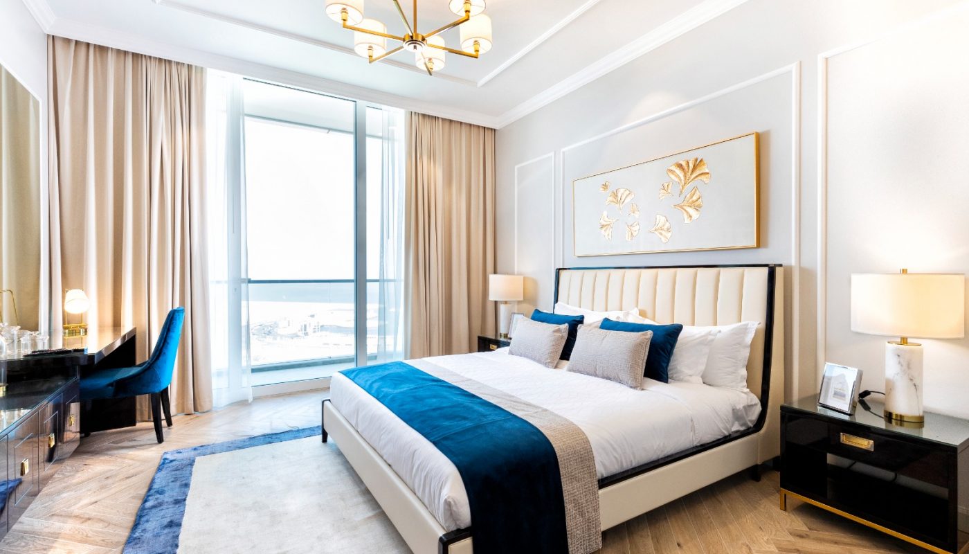 Modern bedroom with a large bed, blue accents, a chandelier, desk with chair, two nightstands, and floor-to-ceiling windows with beige curtains.