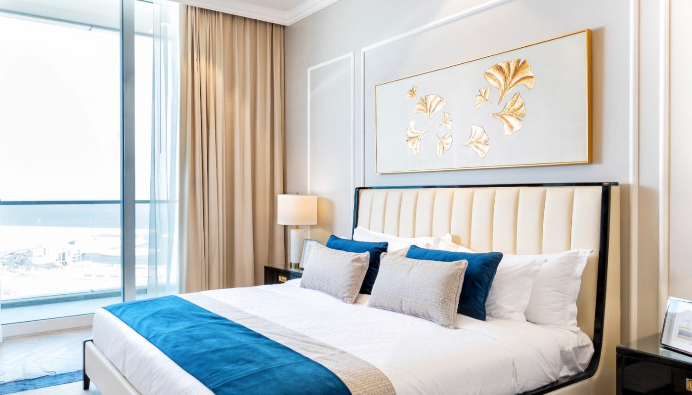 Modern bedroom with a large bed, blue and white bedding, gold wall art, bedside tables with lamps, a window with curtains, and a chandelier overhead.