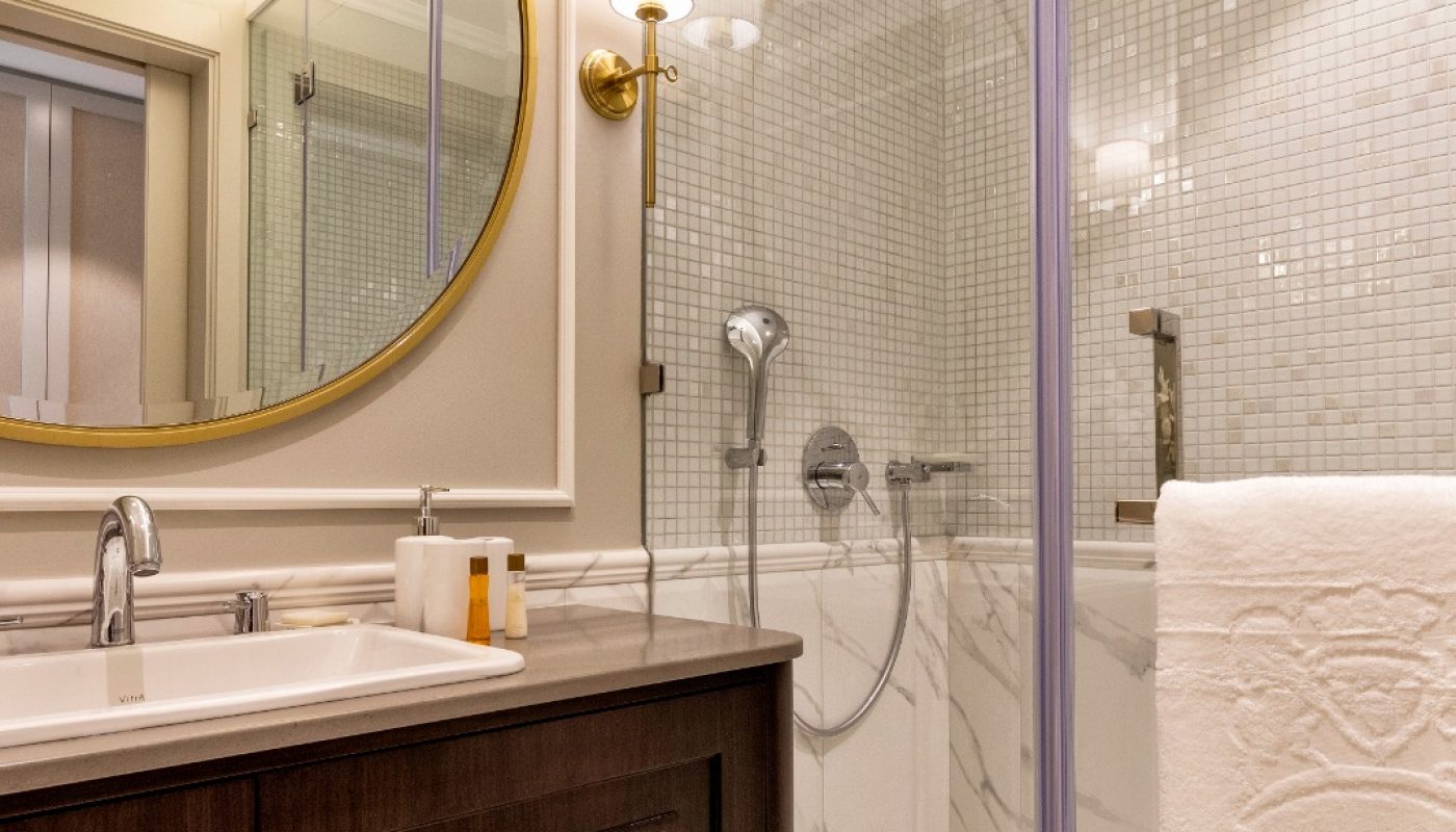 Modern bathroom with a large round mirror, dark wood vanity, marble countertop, and a glass-enclosed shower with white tiles and a towel hanging on the door.