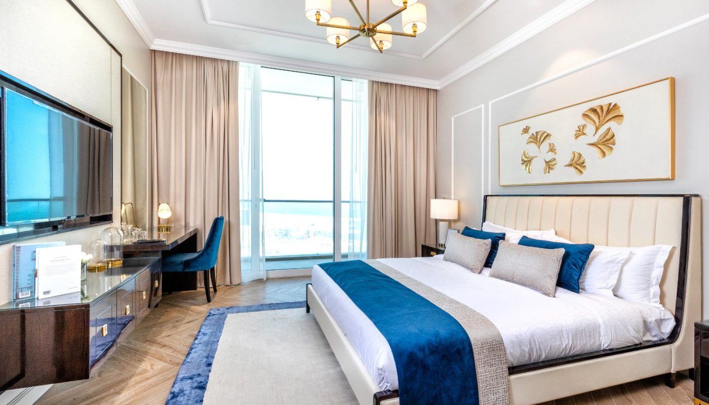 Modern hotel room with a large bed, blue accents, desk, wall-mounted TV, and a window with sheer curtains letting in natural light.