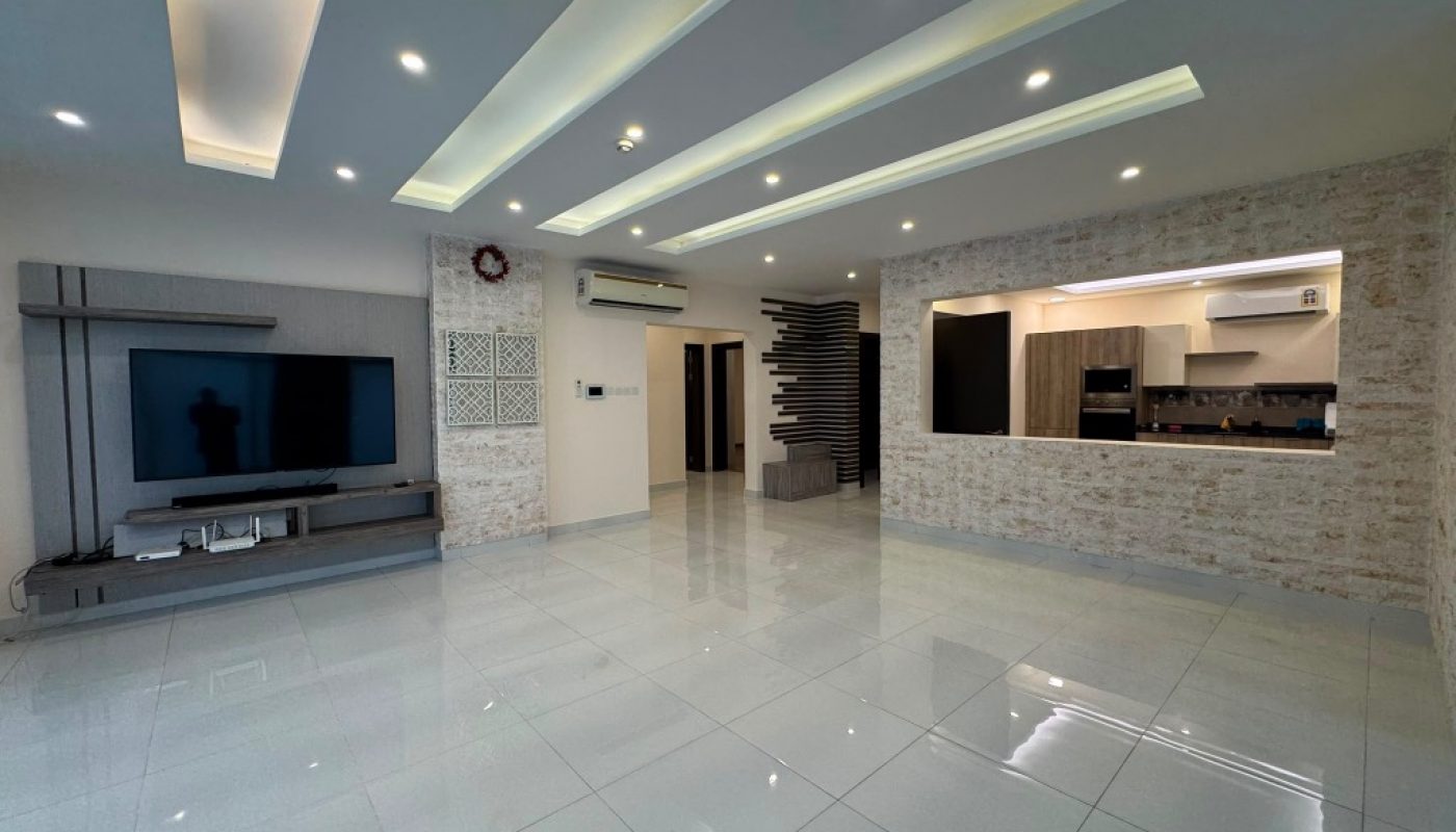 Modern open-plan living room with tiled floors, recessed ceiling lights, a wall-mounted TV, and a partial view of the kitchen area with built-in appliances.