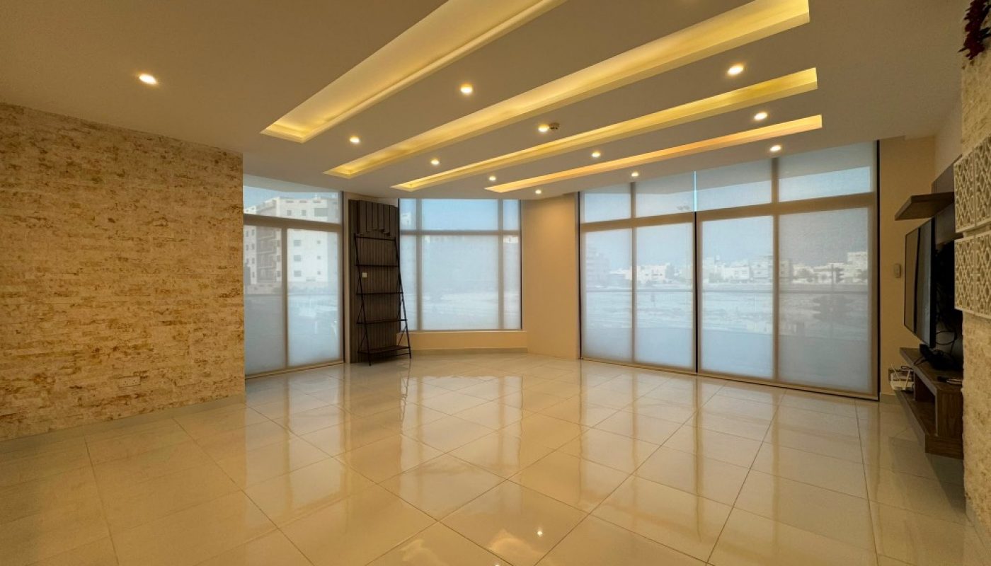 Spacious, empty living room with large windows, tiled floor, recessed ceiling lights, a stone accent wall, and a TV stand in the corner.