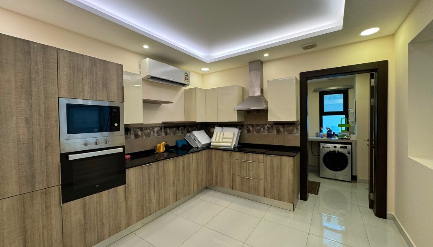 Modern kitchen with wood-finish cabinets, built-in oven, microwave, and stainless steel range hood; laundry area with washer visible through doorway.
