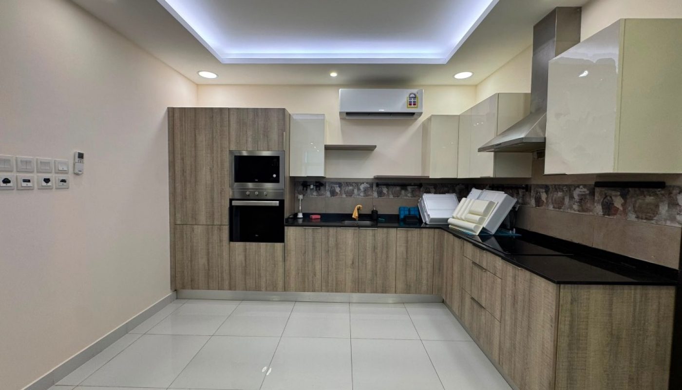 Modern kitchen with wood cabinets, white tiled floor, built-in oven, electric cooktop, range hood, and countertop appliances under soft ceiling lights.