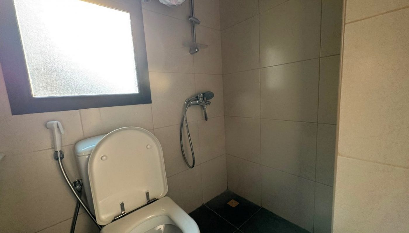 A bathroom with a toilet, handheld bidet sprayer, wall-mounted shower, and tiled floor and walls. Sunlight enters through a frosted window.